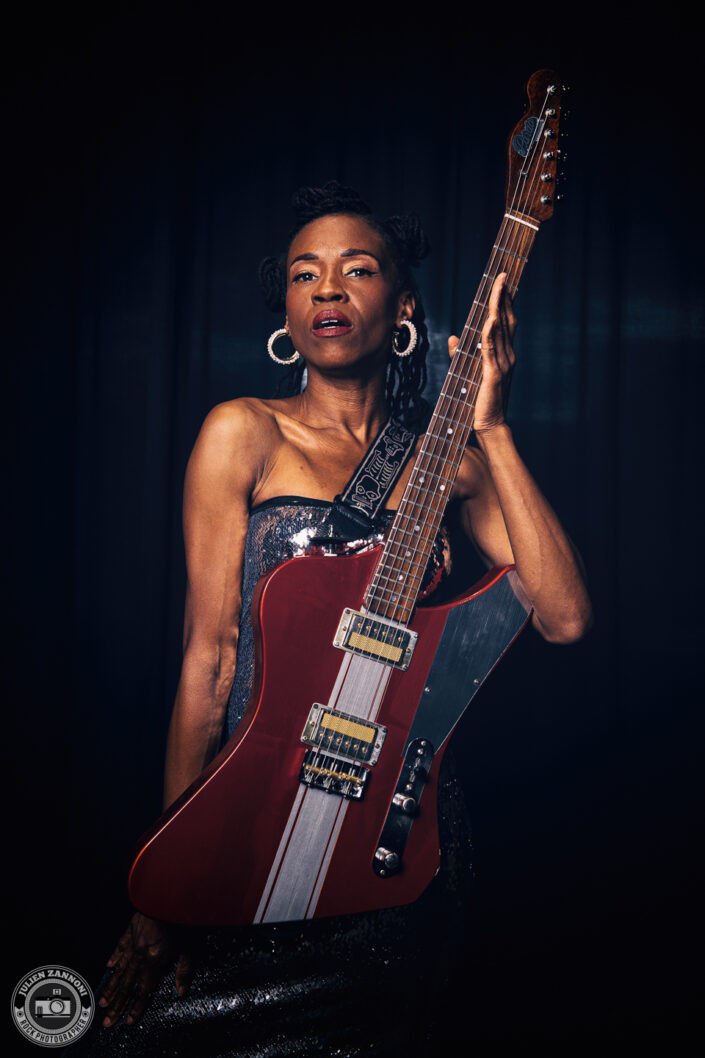 Teneia Sanders performing with her electric guitar in a red dress – photo by Julien Zannoni.