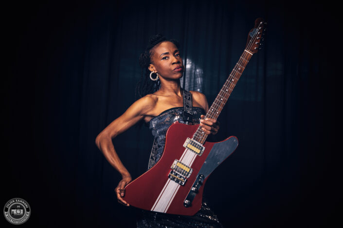 Teneia Sanders posing with her electric guitar against a dark background – photo by Julien Zannoni.