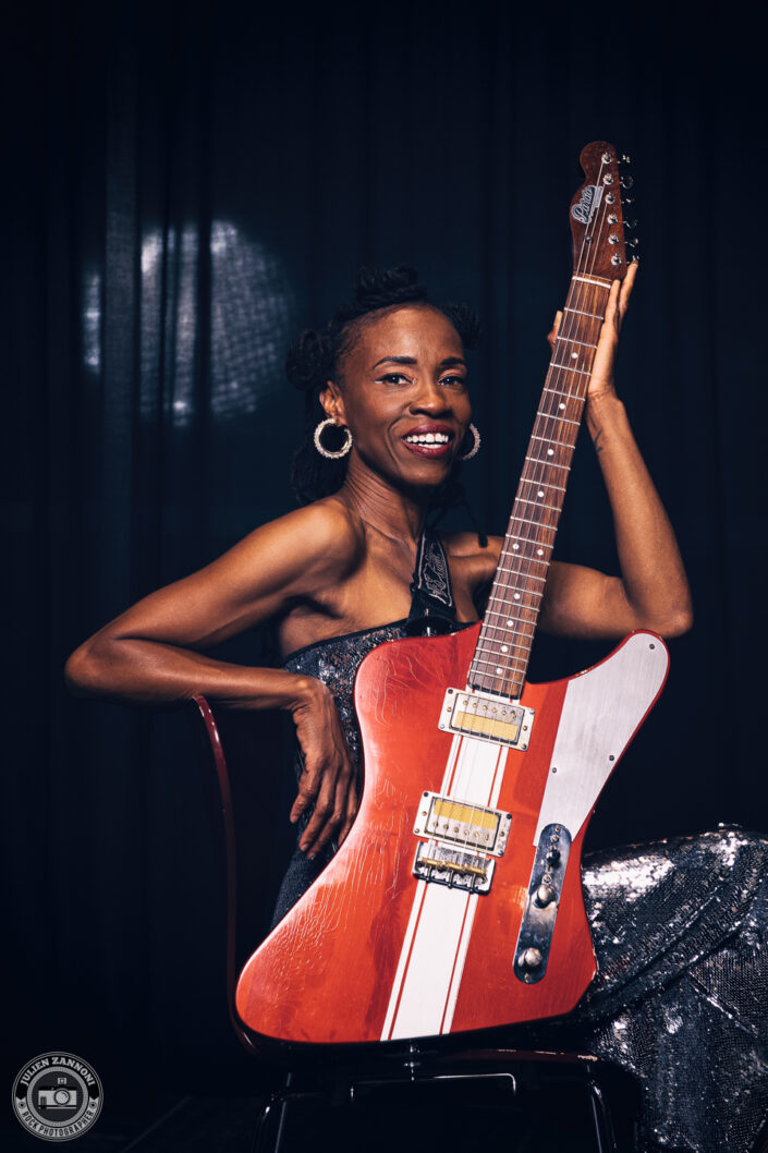 Teneia Sanders smiling while posing with her electric guitar – photo by Julien Zannoni.