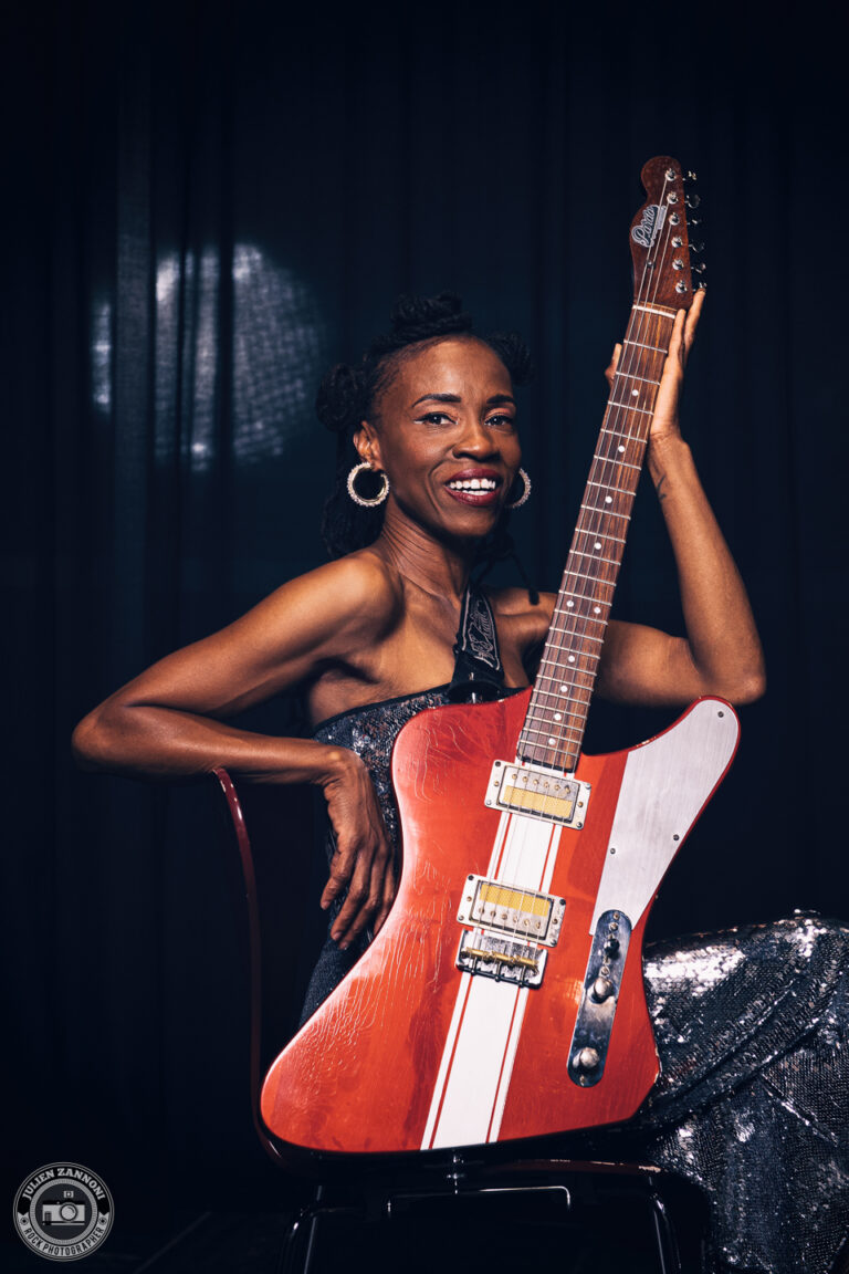 Teneia Sanders smiling while posing with her electric guitar – photo by Julien Zannoni.