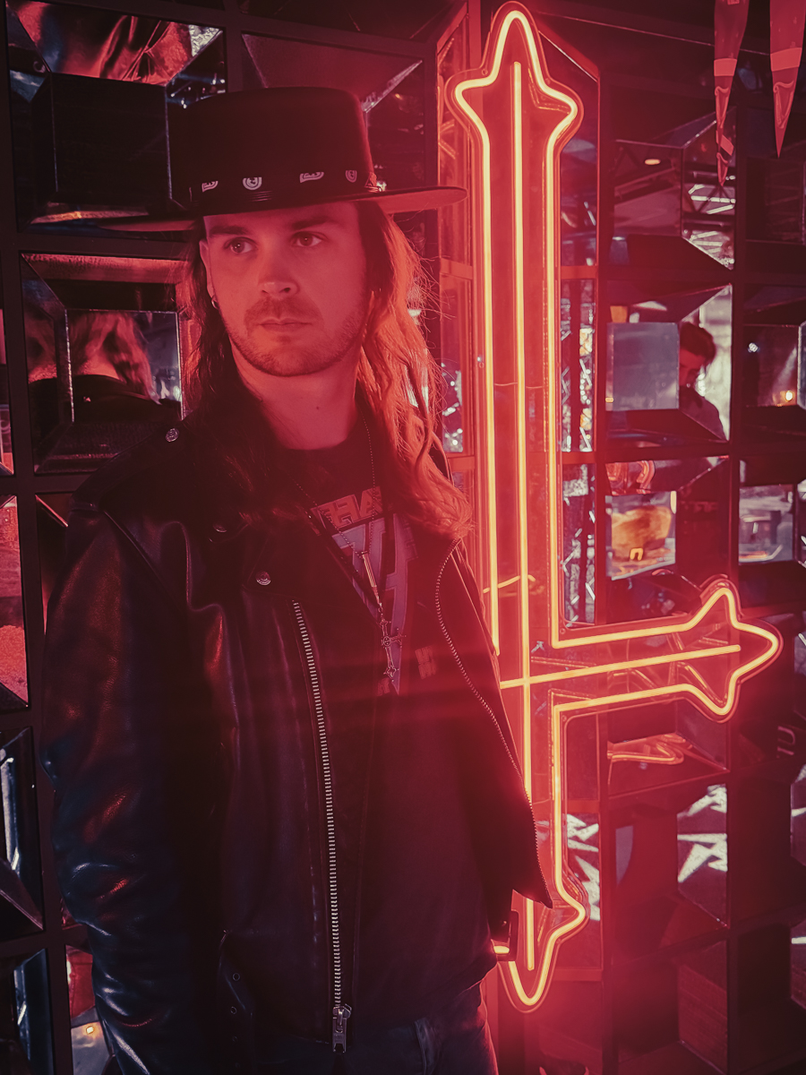 Portrait de Julien Zannoni, photographe, chapeau noir et veste en cuir, devant une croix inversée en néon rouge au bar Le Saint-Rock à Sévrier
